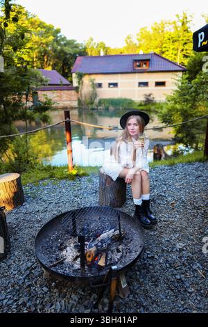 Viaggiatrice che brinda con vino vicino al falò, vestita di bianco e indossa un cappello. Foto Stock