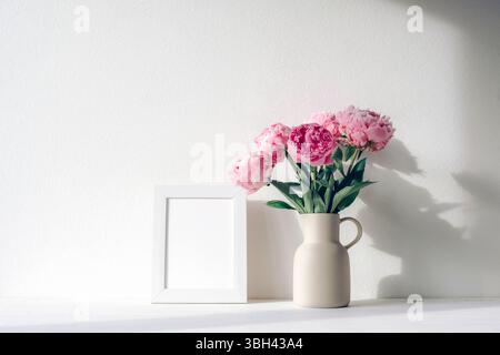 Peonie rosa in caraffa in ceramica e cornice bianca su tavolo bianco con ombra in luce naturale. Mockup. Foto Stock