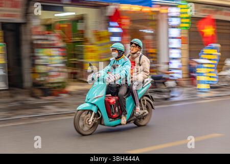 Hanoi, Vietnam - 6 febbraio 2025: Scooter a motore che si muove velocemente con il passeggero sul retro attraverso la strada della città con movimento sfocato sullo sfondo. Foto Stock