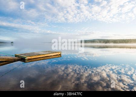 Un molo di legno si estende in un tranquillo lago in Svezia, circondato da nebbie mattutine e morbide nuvole. L'acqua riflette il cielo vibrante, creando un ambiente tranquillo Foto Stock