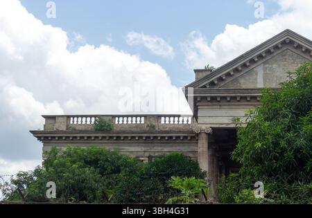 Edificio coloniale in stile neoclassico con decorazioni ornamentali e lussureggiante vegetazione sotto il cielo blu - architettura storica in ambiente tropicale Foto Stock