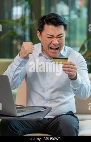 Il giovane e triste ragazzo in camicia seduto nella hall dell'ufficio usando un computer portatile e una carta di credito che si rifiuta di pagare tramite Internet si sente arrabbiato a causa della mancanza di soldi in bancarotta. Concetto di vittima di frode. Foto Stock
