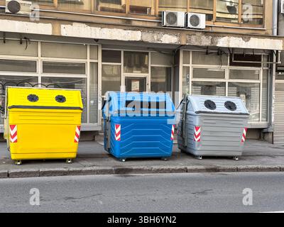 Bidoni di riciclaggio colorati sul marciapiede della città. Contenitori gialli, blu e grigi per la separazione dei rifiuti davanti alla facciata dell'edificio. Foto Stock
