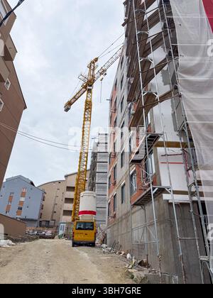 Gru da costruzione alta accanto al blocco di appartamenti incompleto. L'edificio è circondato da impalcature, con materiali da costruzione e una strada sterrata Foto Stock