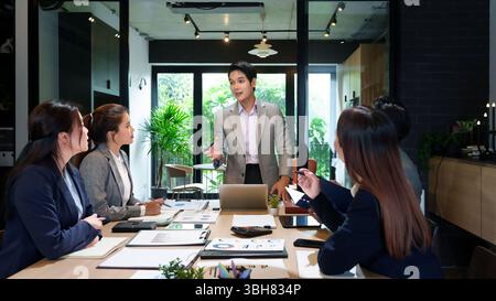 Uomo adulto asiatico che presenta i dati ai colleghi in una riunione. Stanno analizzando i report finanziari e pianificando strategie aziendali in un moderno e luminoso Foto Stock