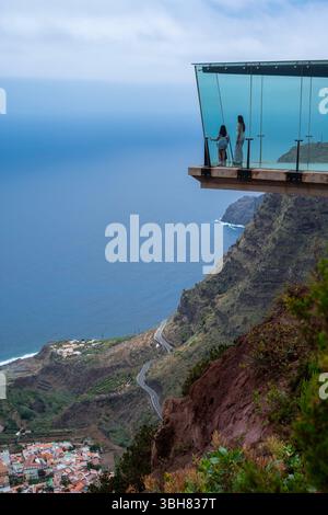 Una coppia si trova su un ponte di vetro che si affaccia sull'oceano. La vista è mozzafiato e la coppia si sta godendo il momento Foto Stock