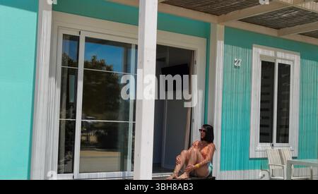Donna rilassante, cabina turchese, libertà estiva: Una giovane donna siede tranquillamente alla porta di una rustica casa sulla spiaggia turchese, che irradia l'atmosfera delle vacanze, sl Foto Stock