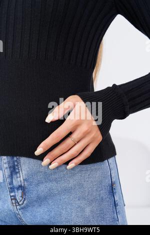 Decorato a mano con un anello delicato si appoggia con grazia sull'anca, indossando un maglione nero a coste. La posa informale e rilassata è completata dal blu chiaro Foto Stock
