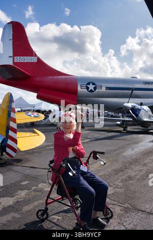 Una donna anziana con una giacca a pois rossa siede in un camminatore di fronte a un grande aereo militare rosso e argento e a un aereo più piccolo. Il set Foto Stock
