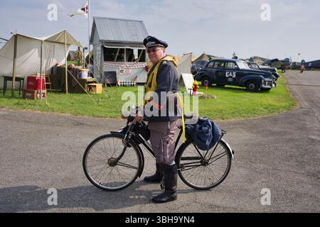 Un uomo in uniforme dell'aeronautica militare tedesca della seconda guerra mondiale si trova con una bicicletta, vicino a tende militari e auto d'epoca a un evento all'aperto. Foto Stock