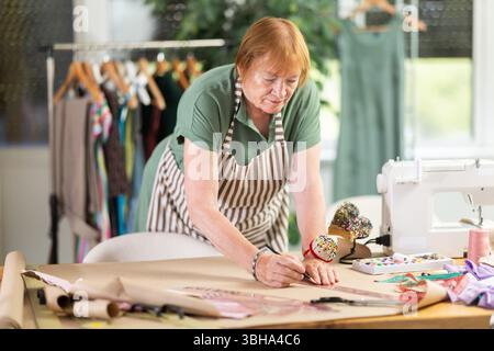 La donna sartoria lavora in un laboratorio di cucito e crea modelli di vestiti su carta. Personalizza con un metro a nastro per creare un design alla moda Foto Stock