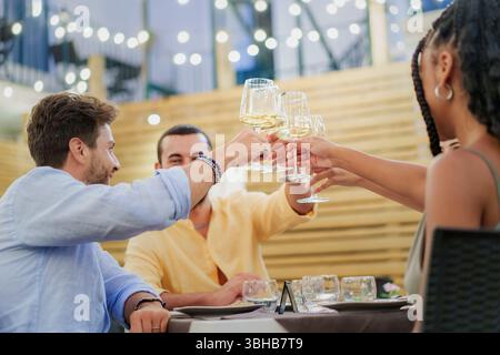 Un gruppo variegato di giovani amici sorride e brinda con vino bianco in un elegante ristorante o bar all'aperto di sera. Una scena di festa Foto Stock