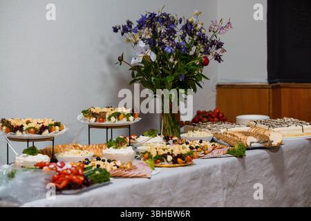 Un tavolo con cibo a buffet ben organizzato presenta un'abbondante varietà di torte, formaggi, frutta e antipasti, completati da una lussureggiante disposizione floreale Foto Stock