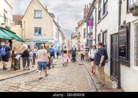 I turisti esplorano l'affascinante Church Street di Whitby, fiancheggiata da pittoreschi negozi ed edifici storici nel cuore della città vecchia. Whitby strada acciottolata. Foto Stock
