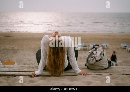 Donna appoggiata sulla panchina della spiaggia, di fronte al tramonto con un'atmosfera tranquilla Foto Stock