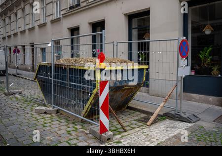 Grande cassonetto metallico riempito di macerie dietro la recinzione metallica protettiva sulla strada della città, Foto Stock