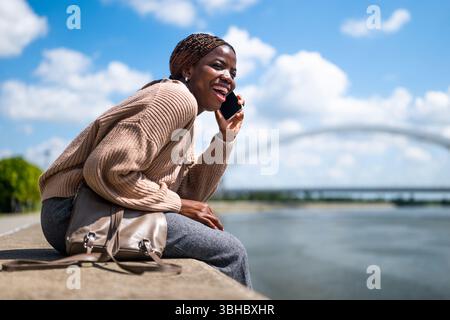 La giovane allegra donna nera sta parlando al telefono. Sta ridendo e si sta godendo la giornata di sole in città. Foto Stock