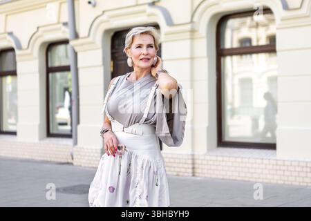 Elegante donna di mezza età con capelli biondi corti in t-shirt grigio chiaro e gonna bianca alla moda in posa all'aperto. Ritratti bella modella senior noi Foto Stock