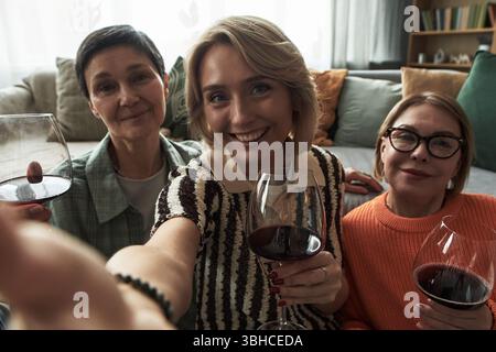 Ritratto di tre donne caucasiche adulte sorridenti e con bicchieri di vino rosso sedute vicino sul divano, amici di mezza età che si divertono insieme e guardano la macchina fotografica Foto Stock