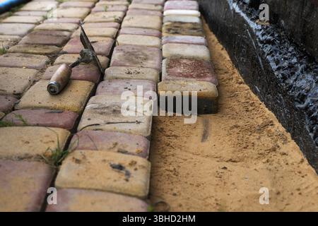 Il lavoratore installa mattoni di pavimentazione sulla sabbia accanto alle fondamenta nere impermeabilizzate. Primo piano del processo di costruzione e di architettura paesaggistica. Foto Stock