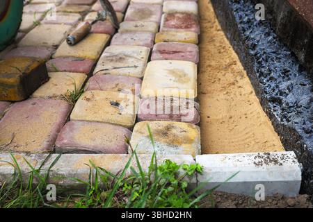 Il lavoratore installa mattoni di pavimentazione sulla sabbia accanto alle fondamenta nere impermeabilizzate. Primo piano del processo di costruzione e di architettura paesaggistica. Foto Stock