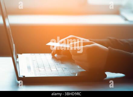 Un uomo tiene una carta bancaria in mano e digita su una tastiera di un portatile, paga gli acquisti, effettua online banking attraverso un'applicazione, vista ravvicinata di Foto Stock