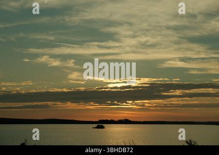 Una barca solitaria galleggia su un lago tranquillo durante un tramonto mite. Strati di nuvole scure e dorate si estendono attraverso il cielo, generando riflessi. Foto Stock