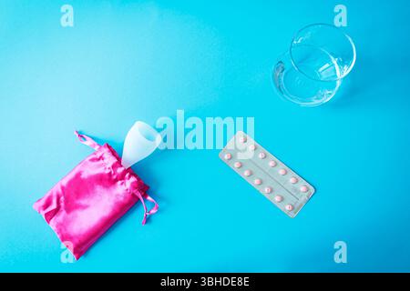 Tazza mestruale in busta rosa, pillole anticoncezionali e bicchiere d'acqua su sfondo blu. Flat Lay circa la salute femminile, la contraccezione, e peri moderni Foto Stock