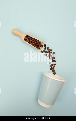 Chicchi di caffè che cadono dal paletto al bicchiere di carta su sfondo blu, vista dall'alto. Posizione piatta. Foto Stock