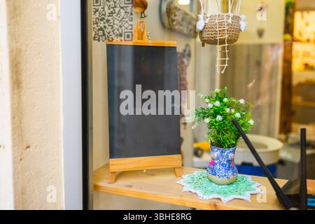 Cartello lavagna vuota con piccola pianta nella finestra davanti al magazzino. Allestimenti creativi, atmosfera accogliente e spazio per messaggi personalizzabili. Foto Stock