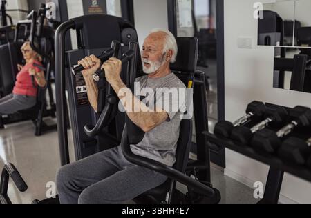 I partner anziani della palestra che utilizzano macchine a mosca per allenarsi, si concentrano sull'anziano che utilizza le attrezzature con sicurezza e facilità Foto Stock