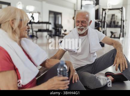 Coppia di anziani seduti sul pavimento e riposati dopo un intenso allenamento in palestra, bottiglia d'acqua in mano e asciugamani intorno alle spalle Foto Stock