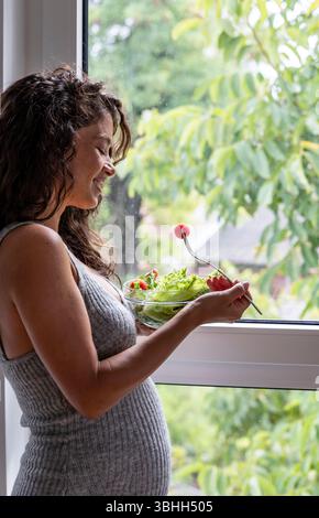Una donna incinta sorridente mentre si trova davanti a una finestra chiusa e mangia un'insalata di verdure per colazione Foto Stock