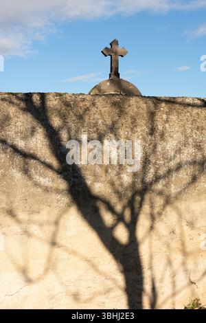 Una suggestiva ombra di un albero si estende attraverso un muro di pietra intemprata, mentre una croce è visibile in cima al muro sullo sfondo di un cimitero. Francia Foto Stock