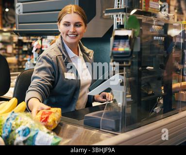 Cassiera sorridente che lavora al registratore di cassa e sposta i prodotti scansionati Foto Stock