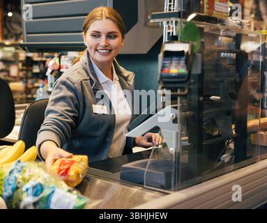 Cassiera sorridente che lavora al registratore di cassa e sposta prodotti scansionati sorridente cassiera che lavora al registratore di cassa e si sposta scansionato Foto Stock