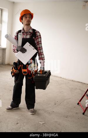 lavoratore maschile all'interno dell'appartamento. Tute da lavoro in piedi in una stanza vuota con cassetta degli attrezzi, appunti e progetti. Riparazioni, const Foto Stock