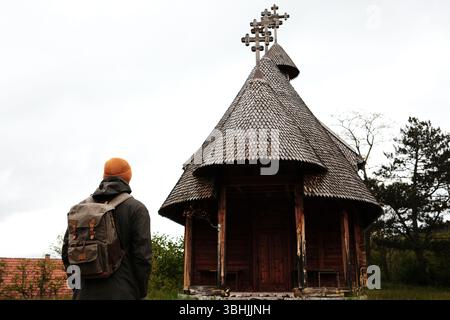 Il viaggiatore maschile con uno zaino si trova di fronte a un'unica chiesa ortodossa in legno con tetto a tegole a Mokra Gora, in Serbia, nella stagione primaverile. Vista posteriore. Foto Stock