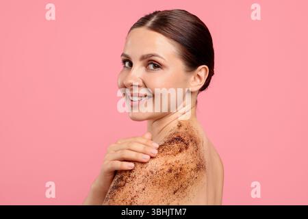 Donna sorridente che applica uno scrub sulla spalla su sfondo rosa Foto Stock