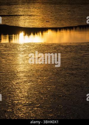 La luce del sole dorata splende sulla superficie ghiacciata di un lago in Svezia. Il sole che tramonta proietta un bagliore arancione sull'acqua ghiacciata e riflette la silhouette Foto Stock