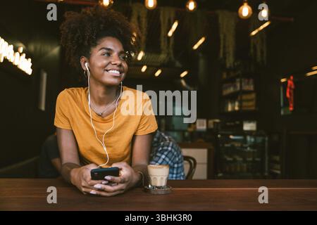 Donna sorridente con auricolari che utilizzano lo smartphone in un accogliente bar e si gusta un caffè Foto Stock
