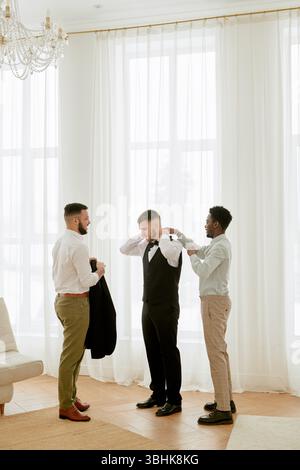 Tre giovani uomini adulti, due caucasici e uno nero, in preparazione alla celebrazione del matrimonio, in piedi insieme in una stanza luminosa, un uomo che regola un altro cravatta mans, terza giacca Foto Stock