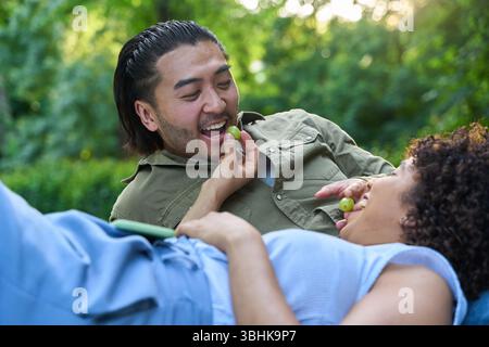 Coppia felice che si diverte durante un romantico picnic in un parco, condividendo l'uva e godendosi il tempo estivo Foto Stock