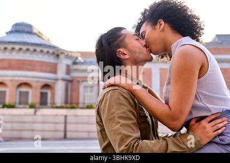 Coppia interrazziale che si baciano appassionatamente nel centro di madrid, con i giardini sabatini sullo sfondo, godendosi un momento romantico durante le loro vacanze Foto Stock