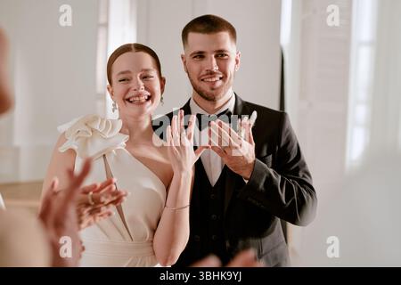 Ritratto di una giovane donna adulta caucasica e di un giovane adulto caucasico che sorride e mostra le fedi di nozze mentre si è in piedi insieme durante la celebrazione del matrimonio, mani alzate, guardando la macchina fotografica Foto Stock