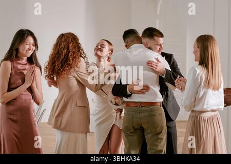 Gruppo di giovani adulti e multietnici che celebrano il matrimonio, sposa che tiene il bouquet sorridente mentre abbraccia lo sposo, amici che si alzano nelle vicinanze applaudono e si congratulano con la coppia Foto Stock