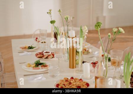 Tavolo per la celebrazione di nozze con stuzzichini assortiti, bicchieri, candele e composizioni floreali, elegante presentazione del cibo su piatti, sedie trasparenti che circondano il tavolo decorato Foto Stock