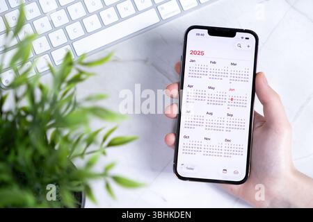 Poznan, Polonia - 10 giugno 2025: Mano con uno smartphone in mostra un calendario completo del 2025 con il nuovo design iOS 26 e Liquid Glass, su una scrivania luminosa Foto Stock