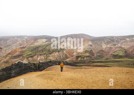 Hiker walks along sand and stone path through the colorful rhyolite mountains of Landmannalaugar in Icelands Highlands, a peaceful journey into wild v Foto Stock