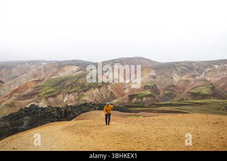 Hiker walks along sand and stone path through the colorful rhyolite mountains of Landmannalaugar in Icelands Highlands, a peaceful journey into wild v Foto Stock